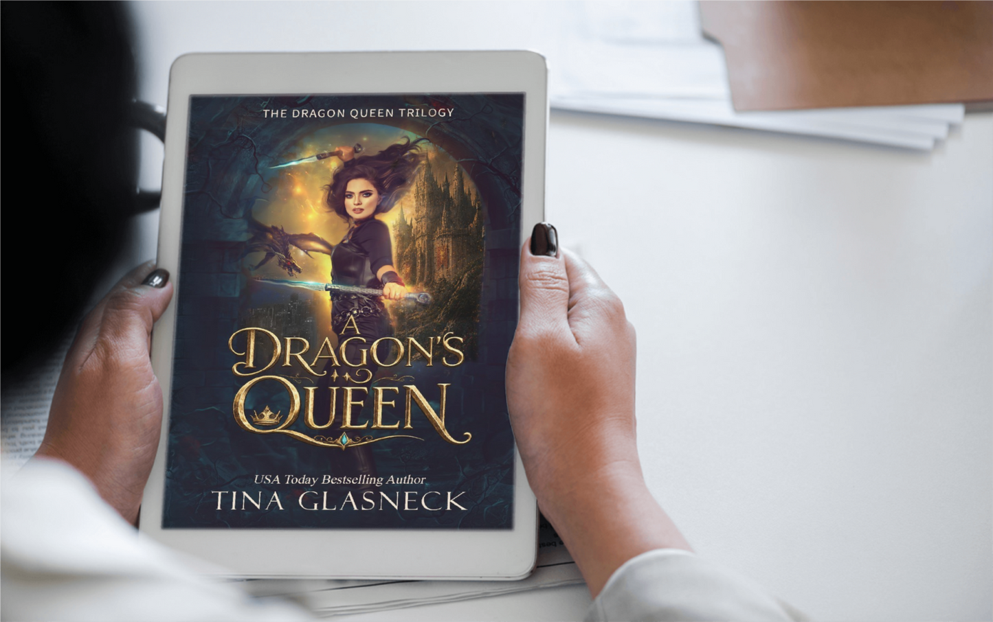A person holds a tablet displaying the book cover of 'The Dragon's Queen' by Tina Glasneck. The cover art features a dark fantasy illustration of a young woman with wings and a glowing dragon, set against a dim, atmospheric background.